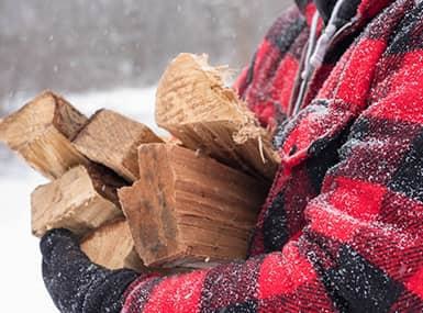 Man wearing a red checkered jacket and black mittens carrying firewood while snowing.