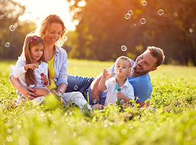 Family of four enjoying spring while blowing soap bubbles in a green field of shrubs and trees.
